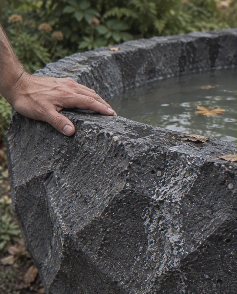 Mão apoiada na borda facetada da banheira de concreto grafite, com folhas secas e água